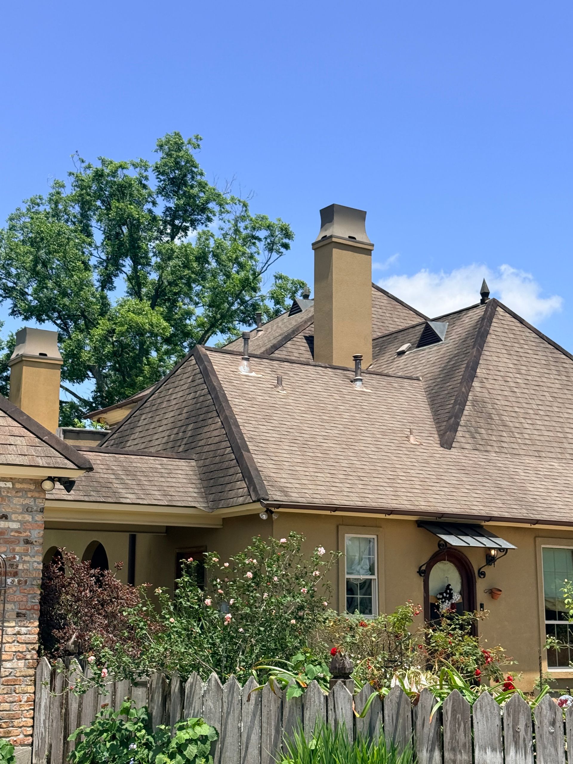 House with tan shingle roof, chimney, and garden under a clear blue sky