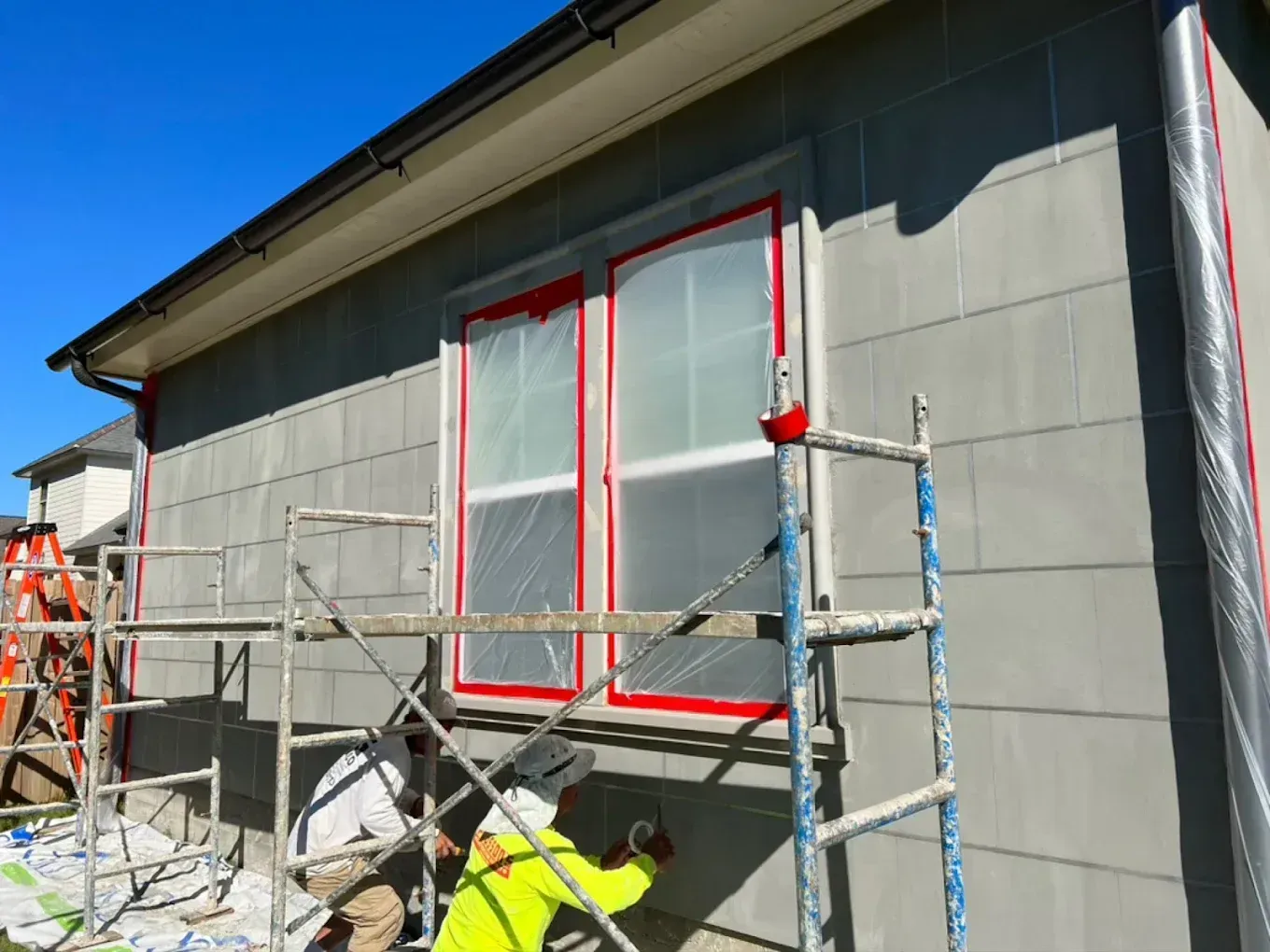 Workers on scaffolding painting a house exterior, with windows masked in red tape.