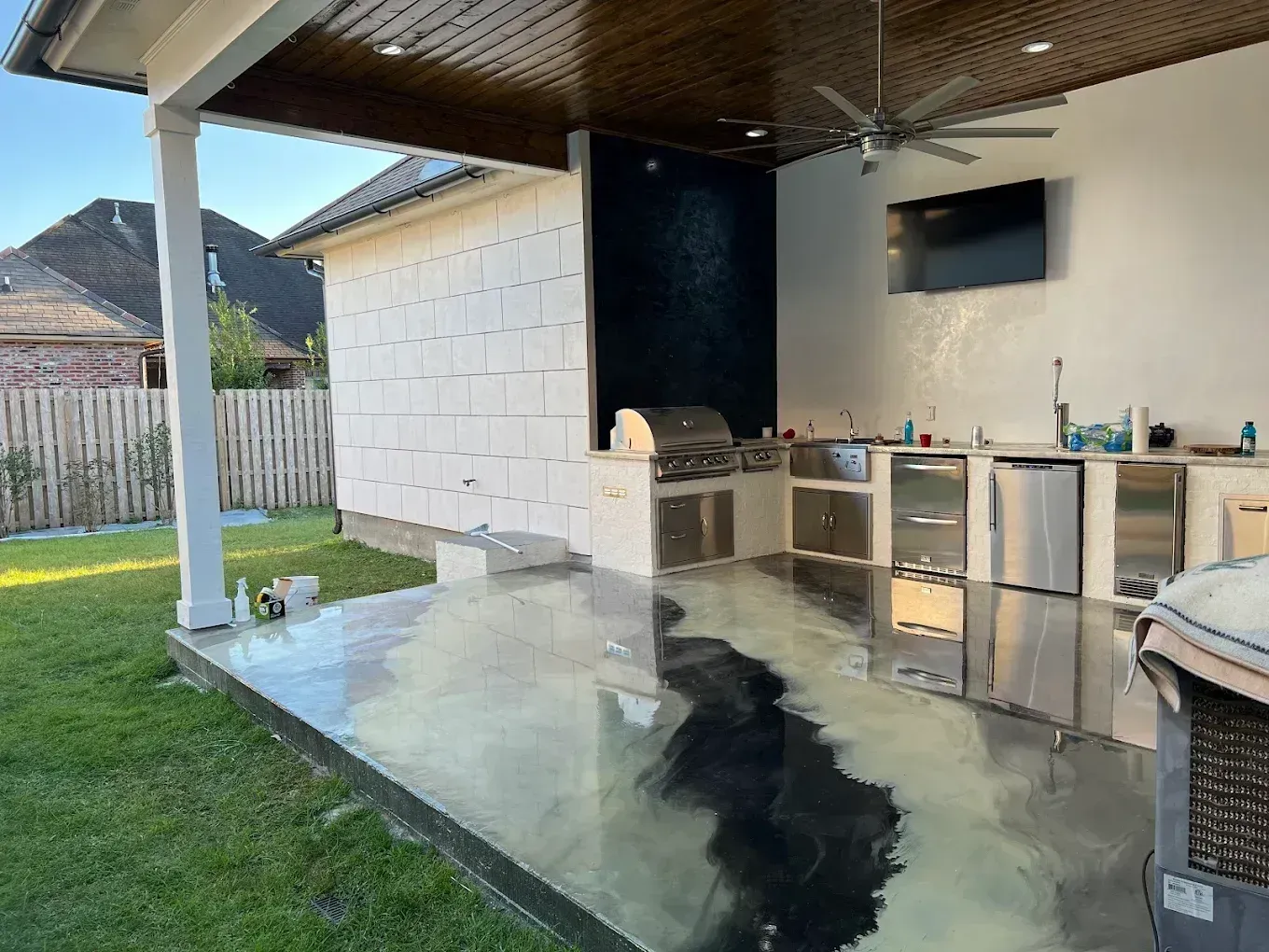 Backyard patio with wet concrete floor, outdoor kitchen cabinets, and a covered roof beside a grassy yard