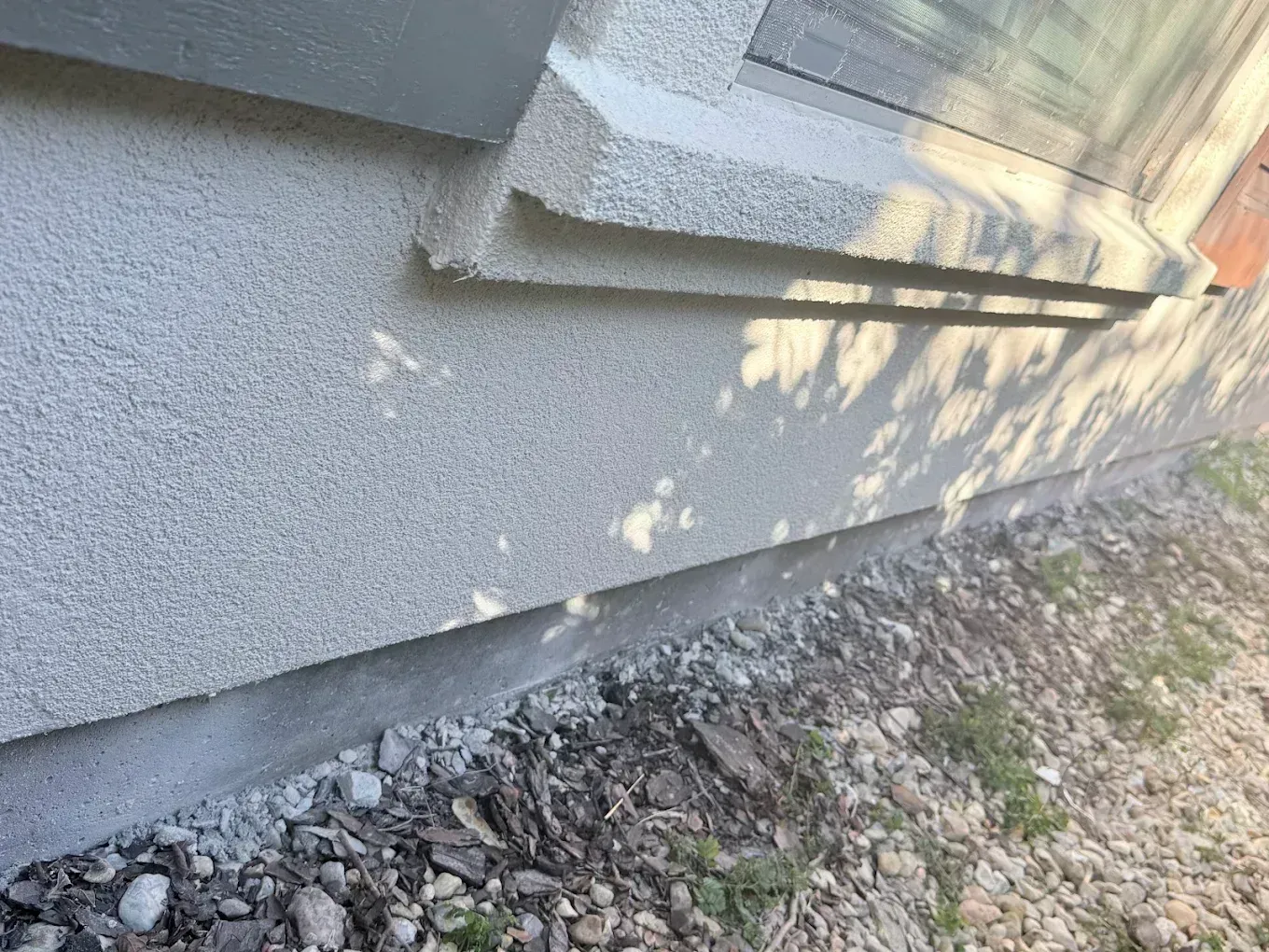 Close-up of a gray stucco wall beside gravel ground and stacked boards in sunlight.
