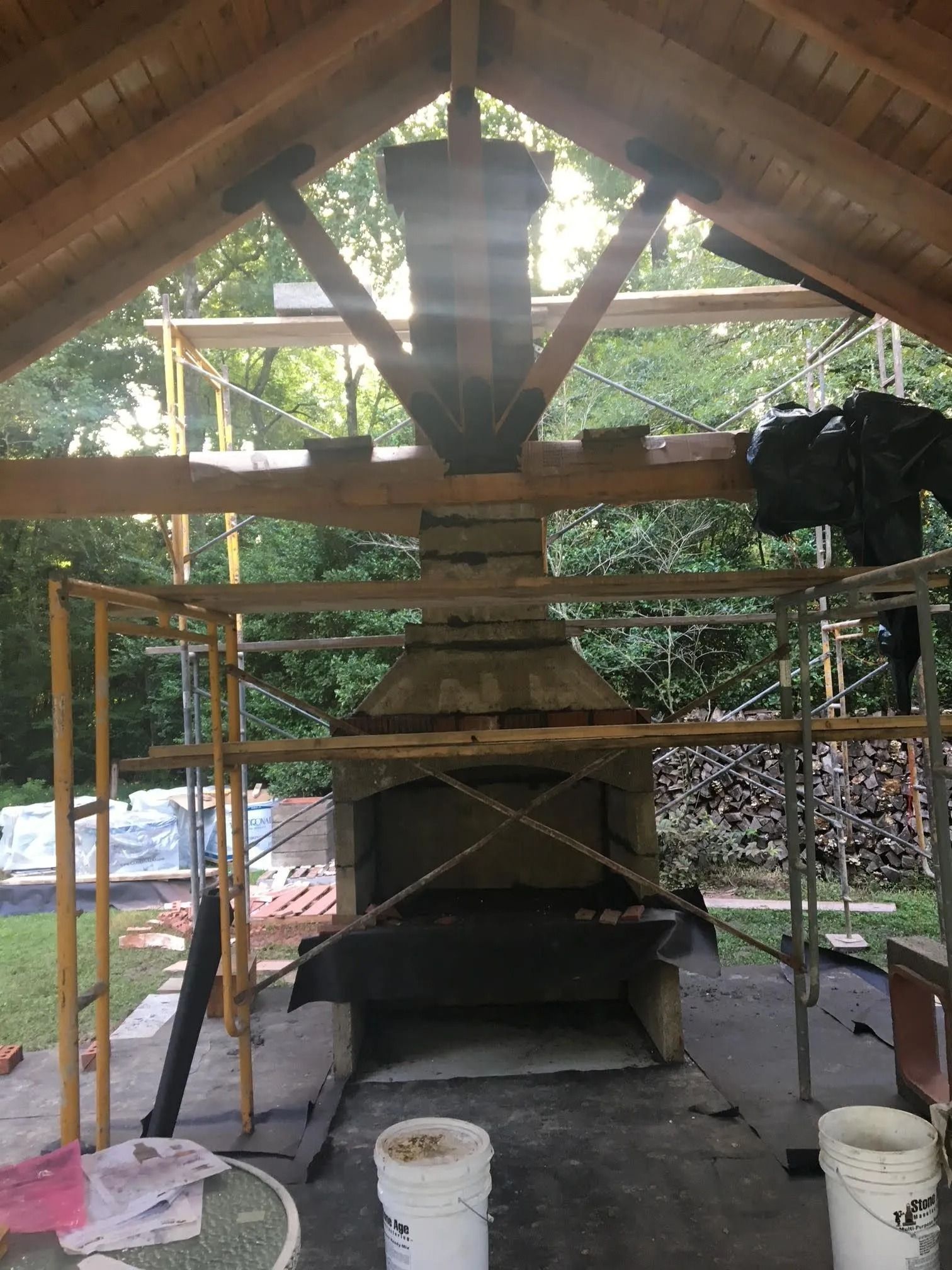 Outdoor wood-fired grill or oven under a wooden shelter, with scaffolding and buckets around it.
