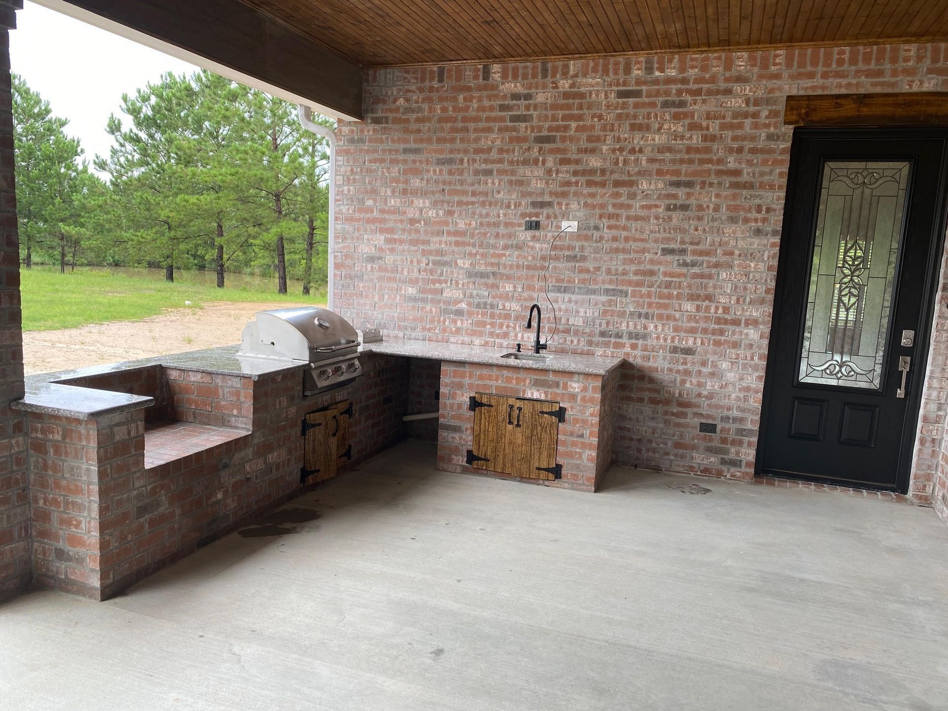 Covered outdoor brick patio with built-in grill, sink, and black door beside a grassy yard