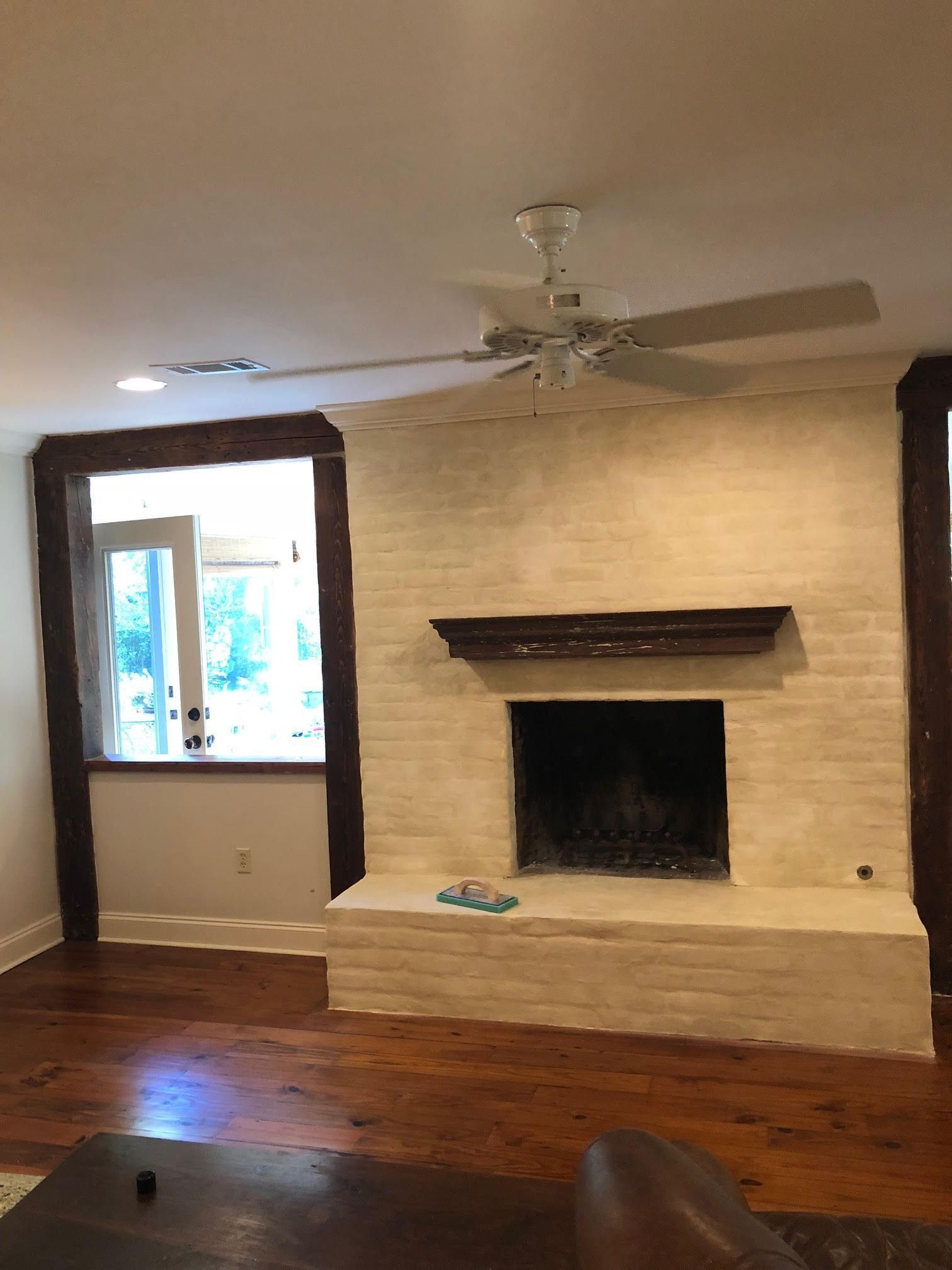 Living room with brick fireplace, hardwood floors, ceiling fan, and a window letting in daylight