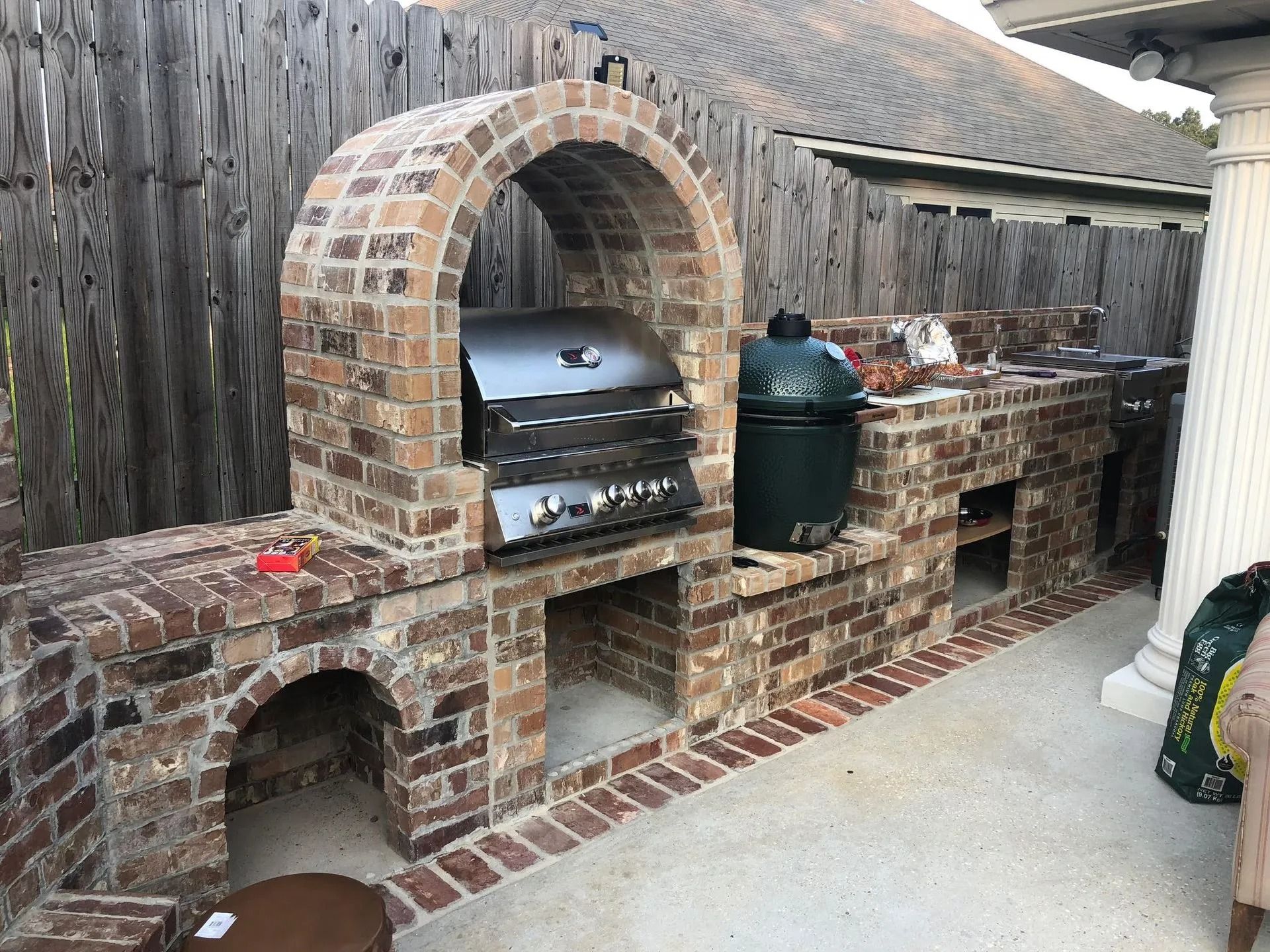 Brick outdoor barbecue grill with arched oven, cooking grates, and a green smoker on a patio