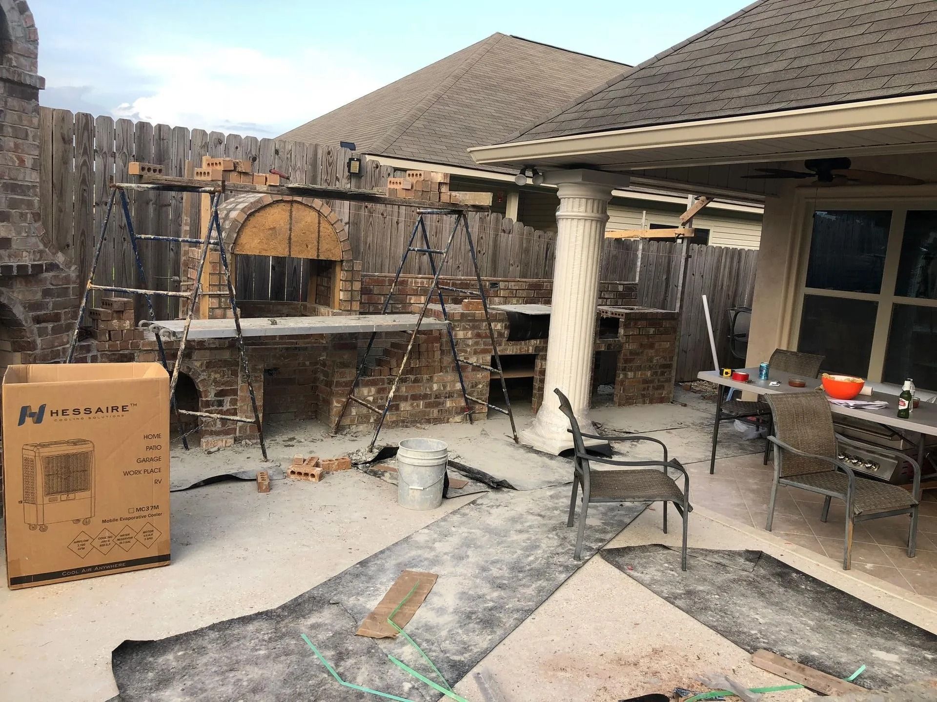 Backyard patio with scattered furniture, stacked firewood, and a cardboard box under a covered pergola