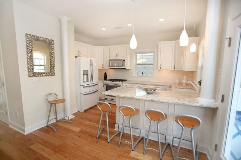 A kitchen with white cabinets , granite counter tops , stools and a refrigerator.