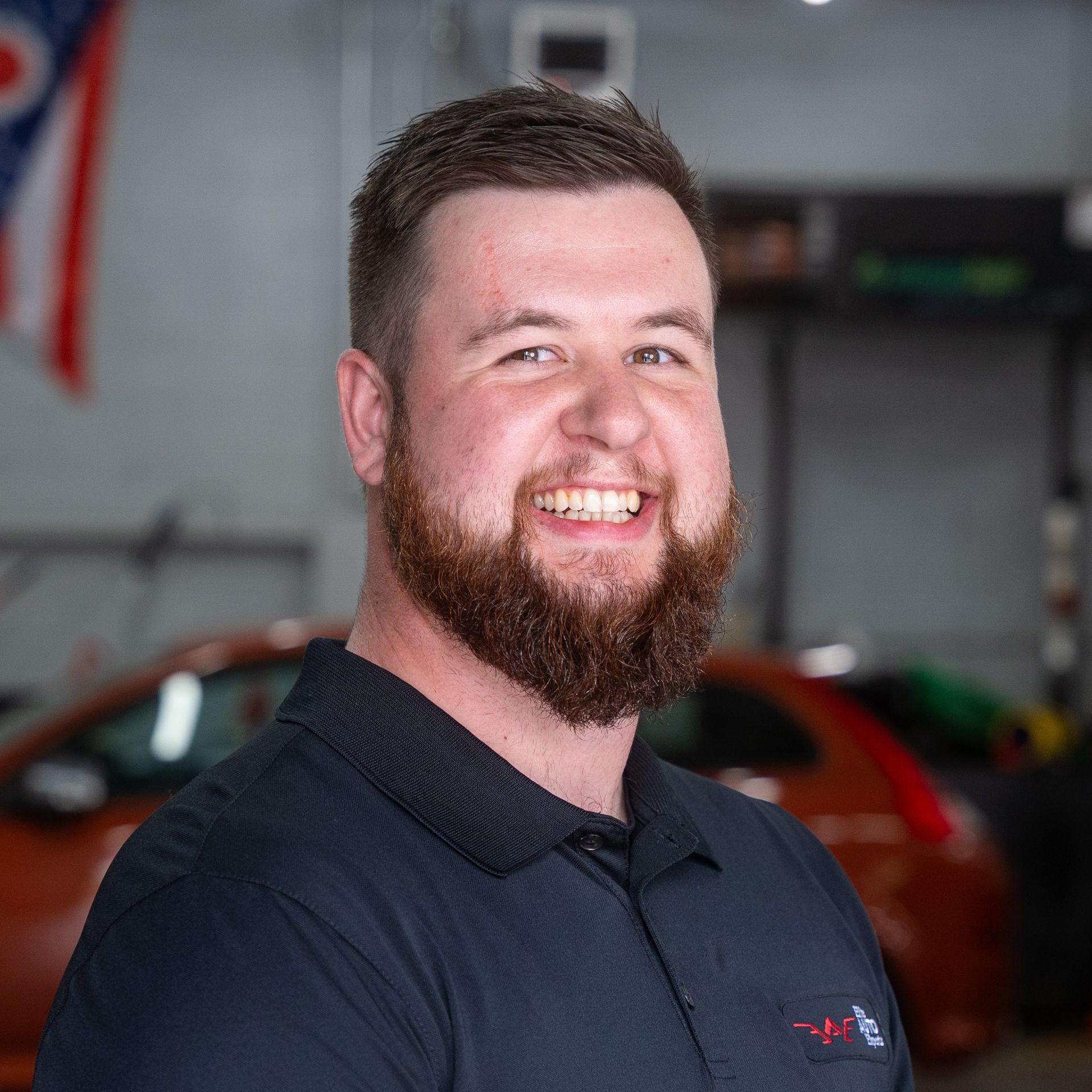 A person with a beard and short hair smiles while wearing a dark polo shirt in a car shop setting with a car in the back.