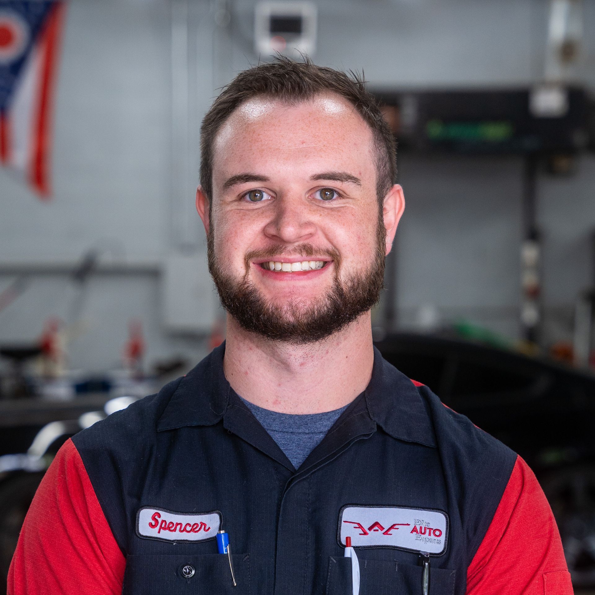 A smiling mechanic in a red and black uniform stands in an automotive workshop.