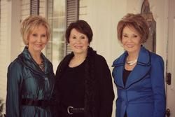 Three women standing indoors, smiling, wearing dark, black, and blue outfits.