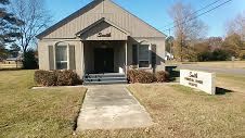 Small beige building with a front walkway and a sign on the right lawn.