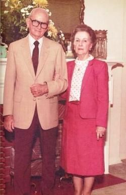 Two people standing indoors, wearing formal clothes, with a fireplace in the background.