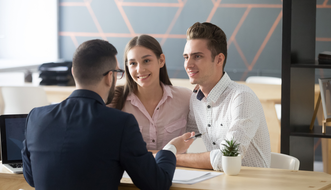 A person in a suit sits across from a smiling couple at a wooden desk in a bright, modern office setting.