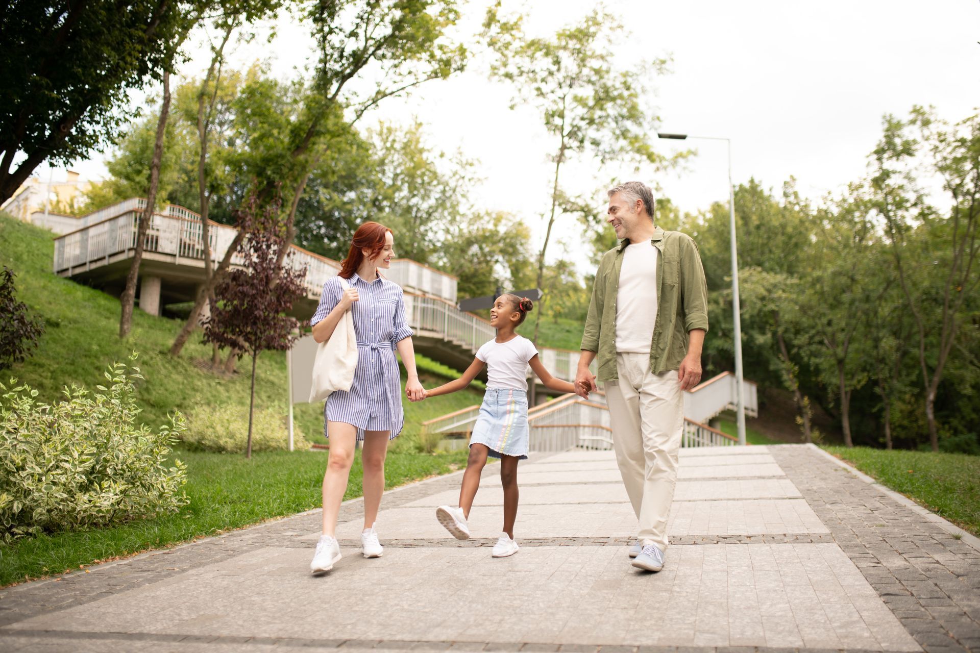 A family walks hand-in-hand along a paved park path, surrounded by green trees and sloping hills.