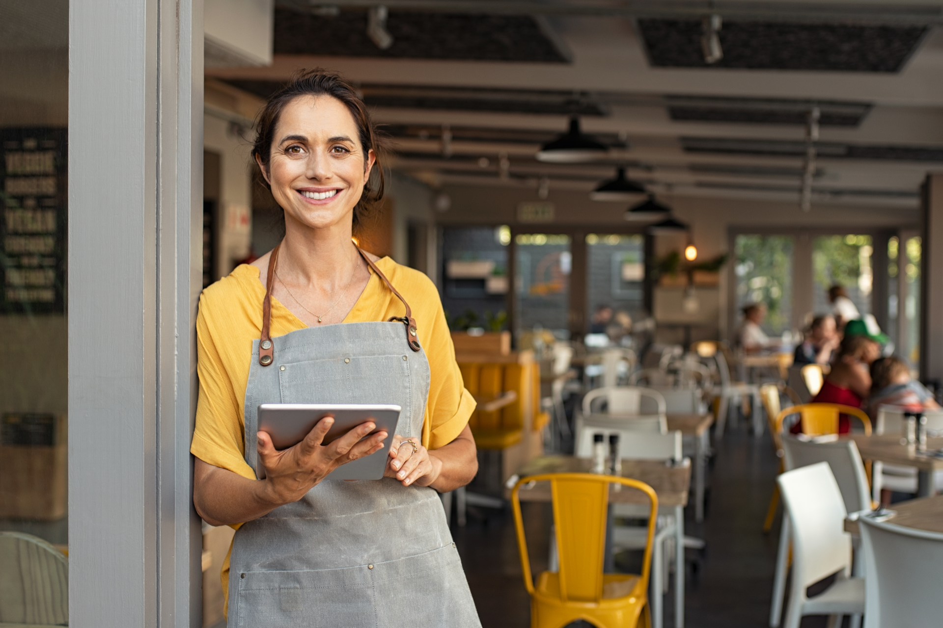 A person wearing a yellow top and grey apron smiles while holding a digital tablet in a restaurant with tables and chairs.