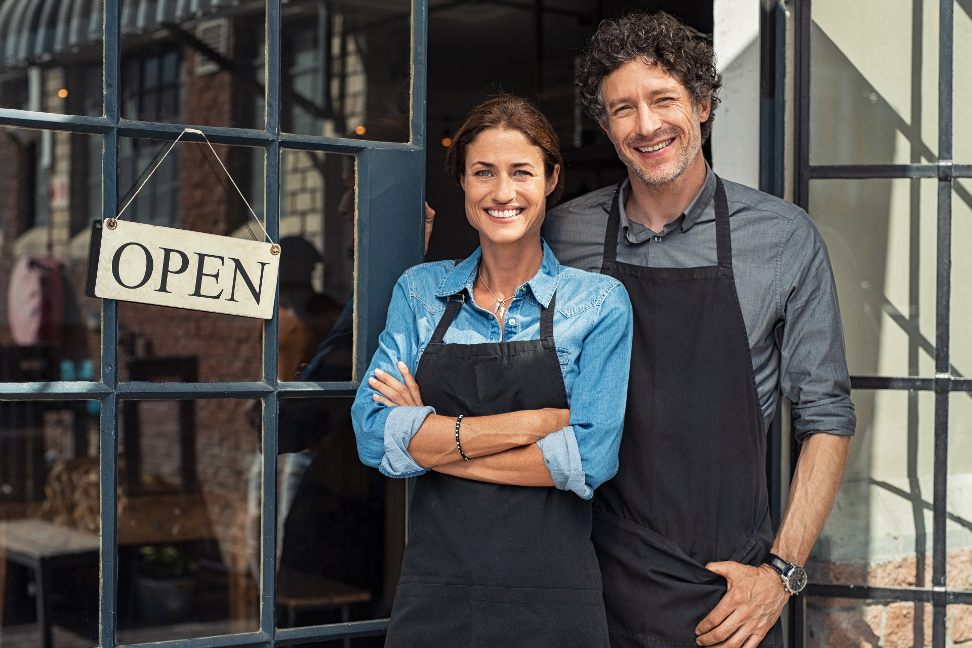 Two people wearing black aprons stand smiling in front of a shop entrance with an