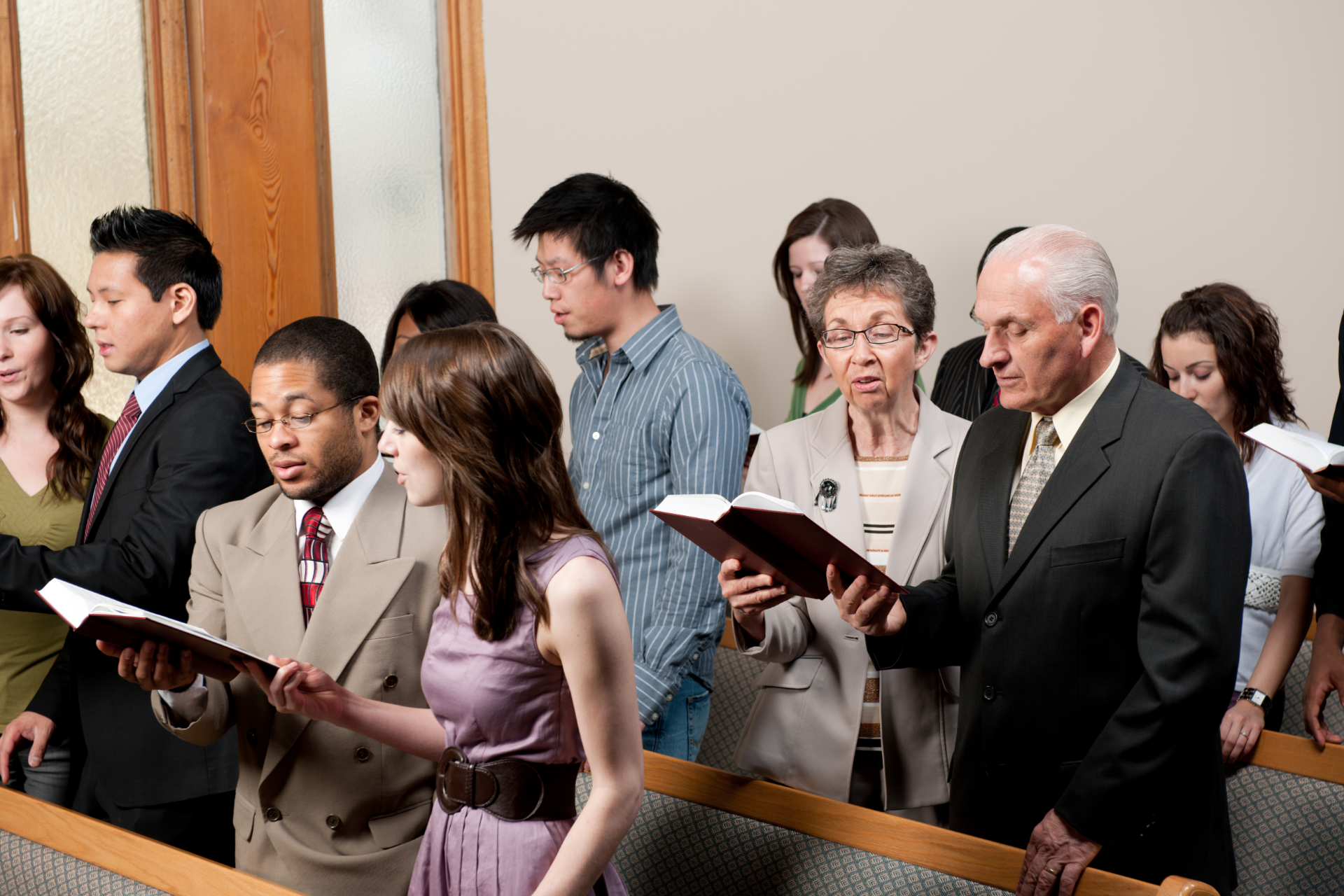 A group of diverse people stand in rows inside a church, holding open hymn books and singing together.
