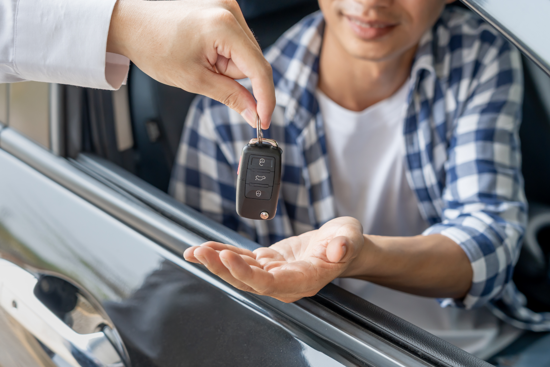 A person hands a car key to another individual sitting in the driver's seat of a vehicle.