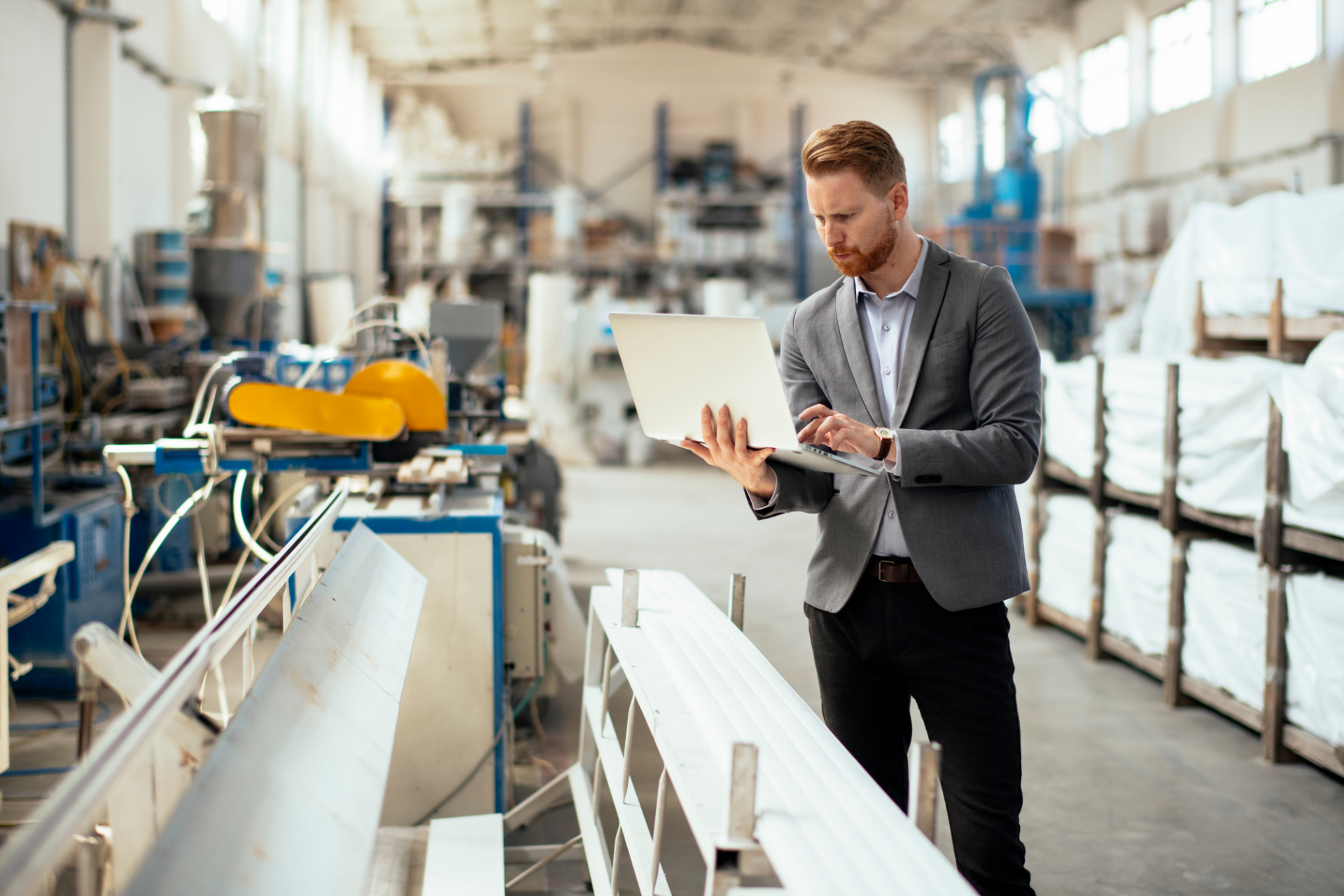 A professional holds a laptop while inspecting industrial machinery in a warehouse with storage racks.
