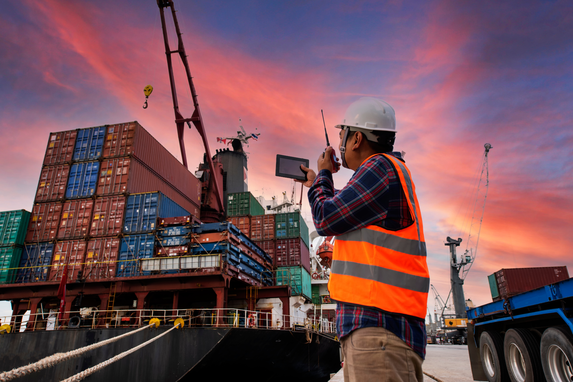 A worker in a safety vest and hard hat uses a radio and tablet near a container ship at sunset.