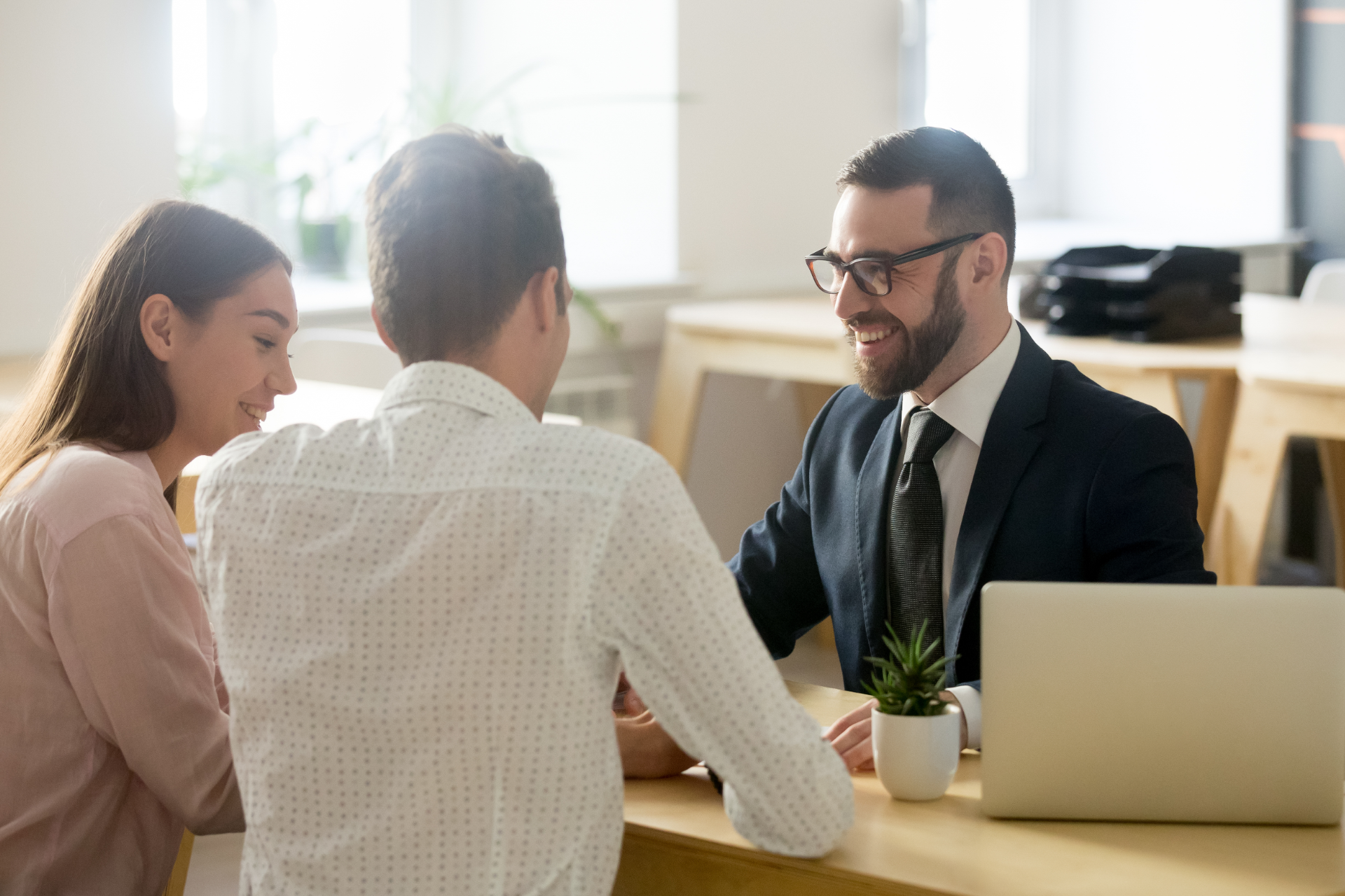 Two people sit at a desk talking to a smiling professional in a suit and glasses in a bright, modern office space.