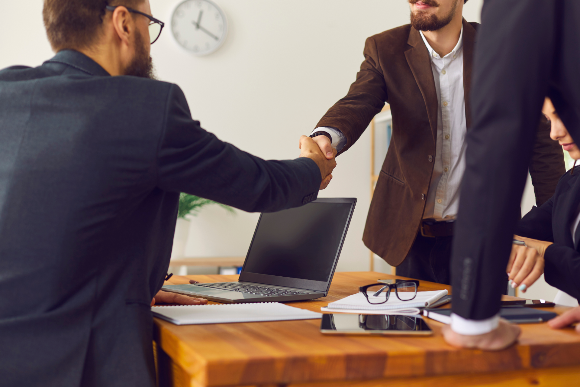 Two business professionals shake hands across a wooden desk in an office setting, with colleagues present during a meeting.