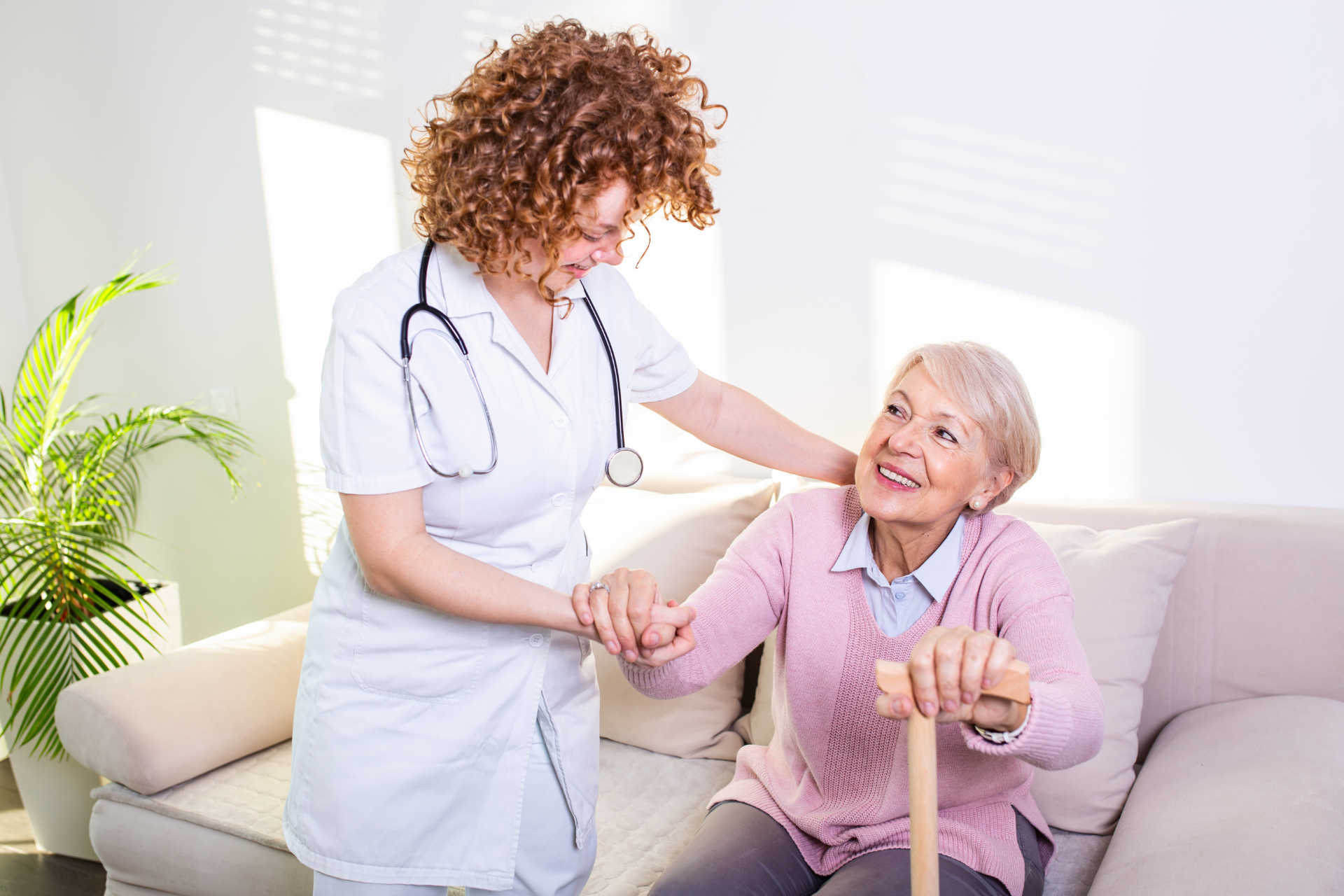 A healthcare worker helps an individual stand up from a sofa using a cane in a bright room.