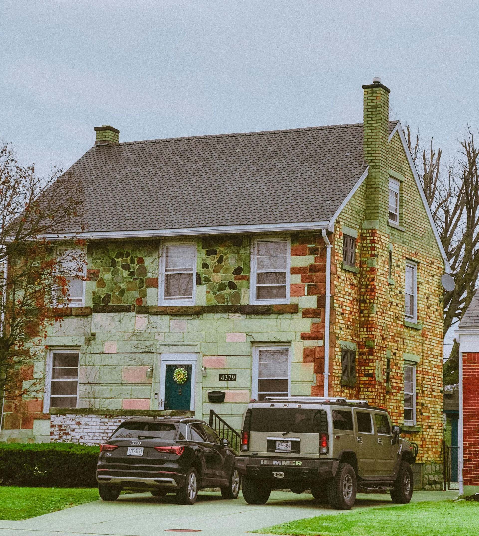 A two-story stone house with a weathered green exterior and a brown shingled roof, with two cars parked in the driveway.