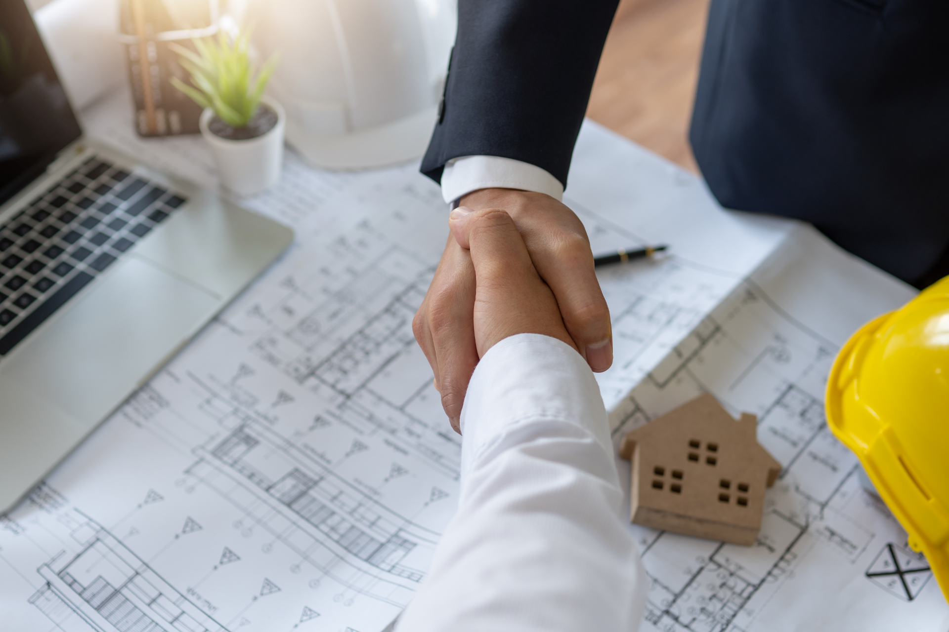 A professional handshake over architectural blueprints with a laptop, miniature house, and yellow hard hat nearby.