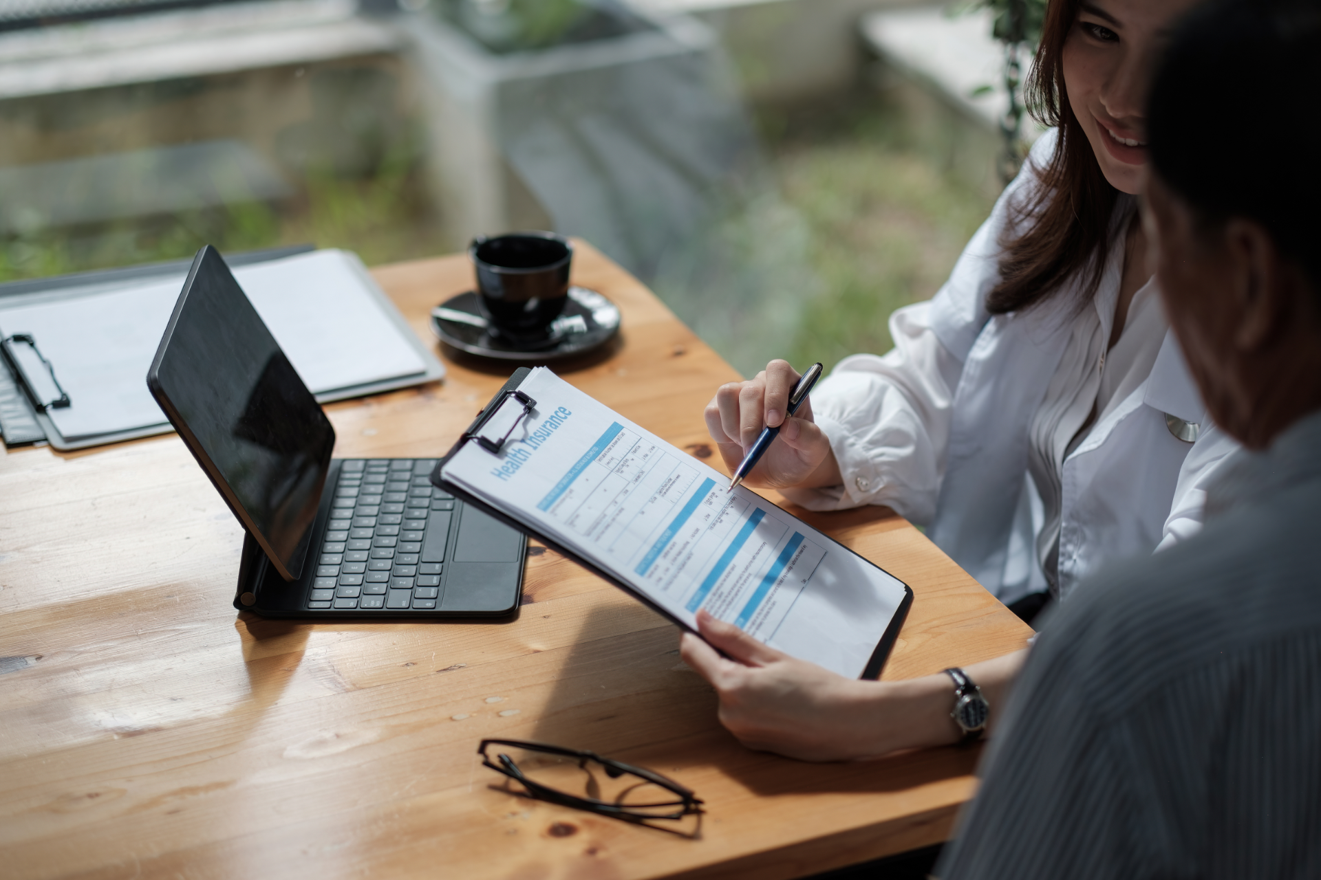 A professional meeting where someone points to a document on a clipboard while seated at a wooden desk with a tablet.