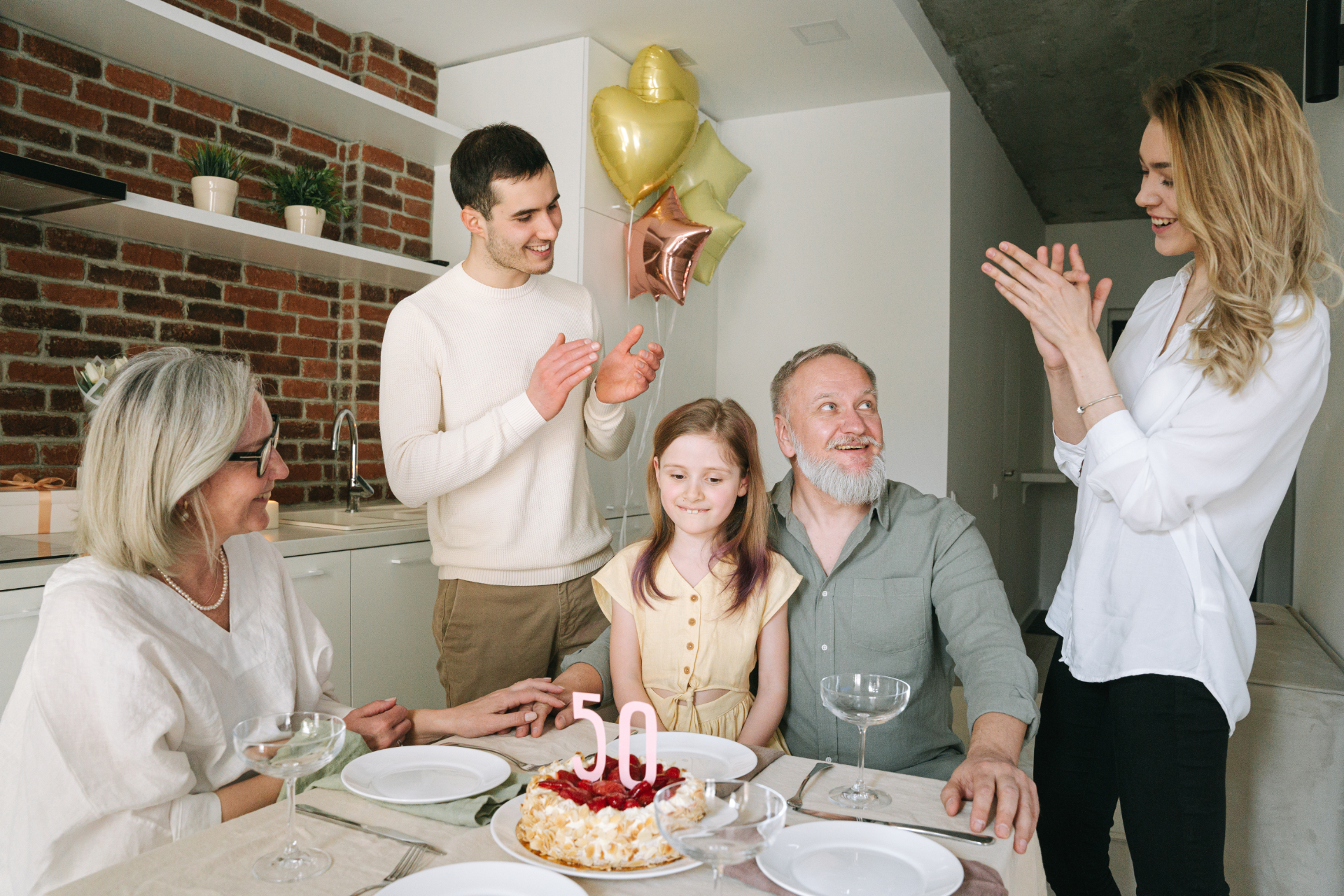 Family outdoors, smiling and talking; man gesturing, elderly man sitting, young girl, and blonde women.