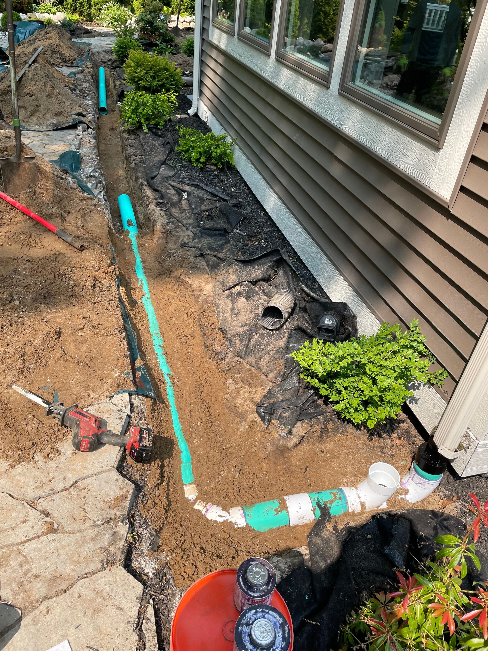 Garden trench with exposed pipes beside a house, bordered by soil, concrete, and a small green shrub.