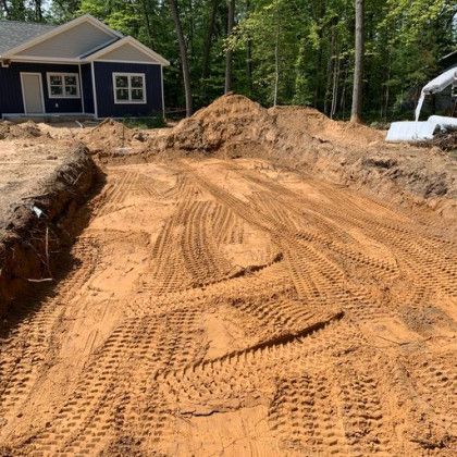 Freshly graded dirt lot beside a blue house and wooded area, with tire tracks and soil piles.