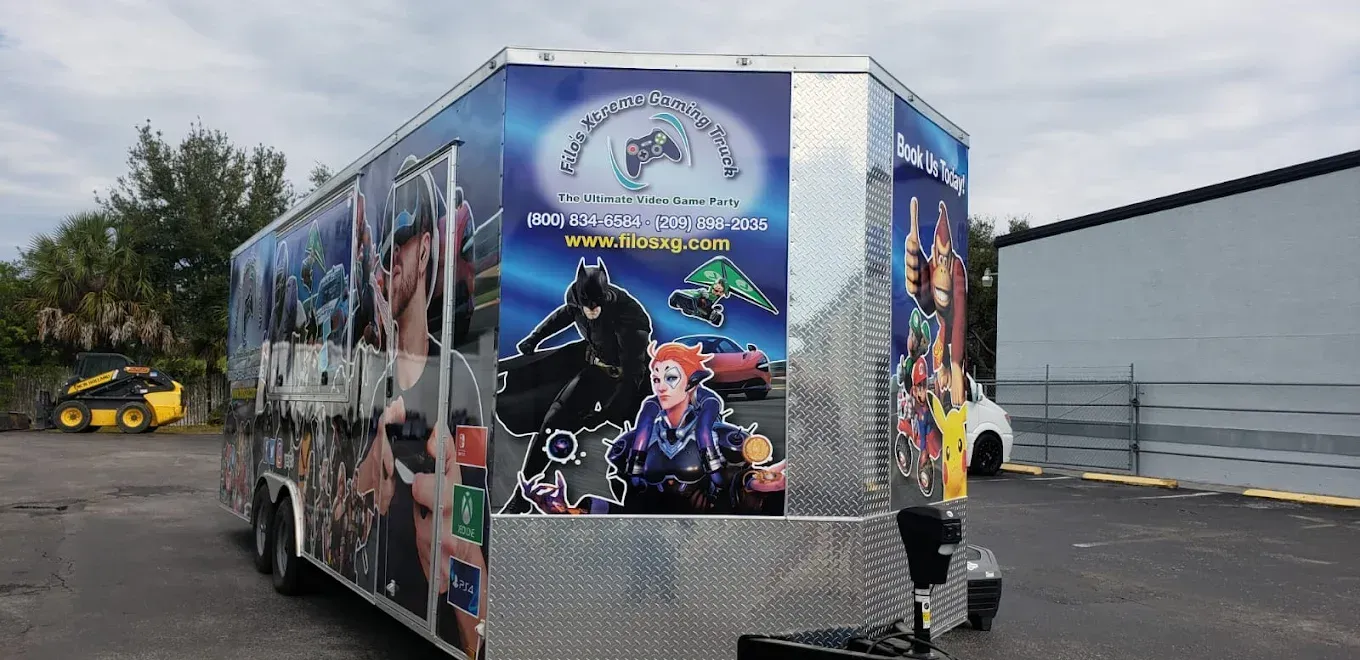 A game truck trailer with comic book characters and a logo against a cloudy sky.
