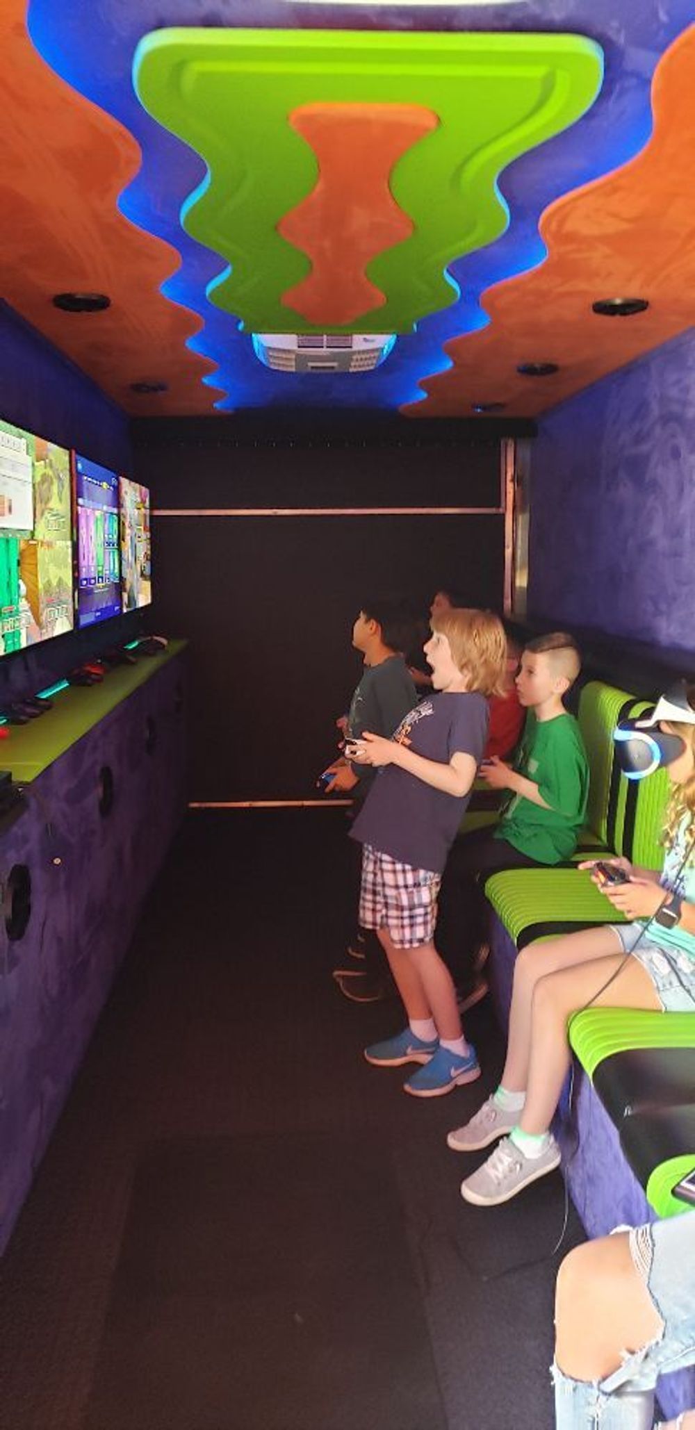 Inside a brightly decorated game truck, kids play video games. The ceiling is orange, green, and blue.