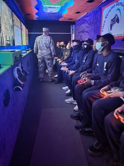 Inside a gaming truck, several people sit playing video games, an uniformed soldier stands in the middle.