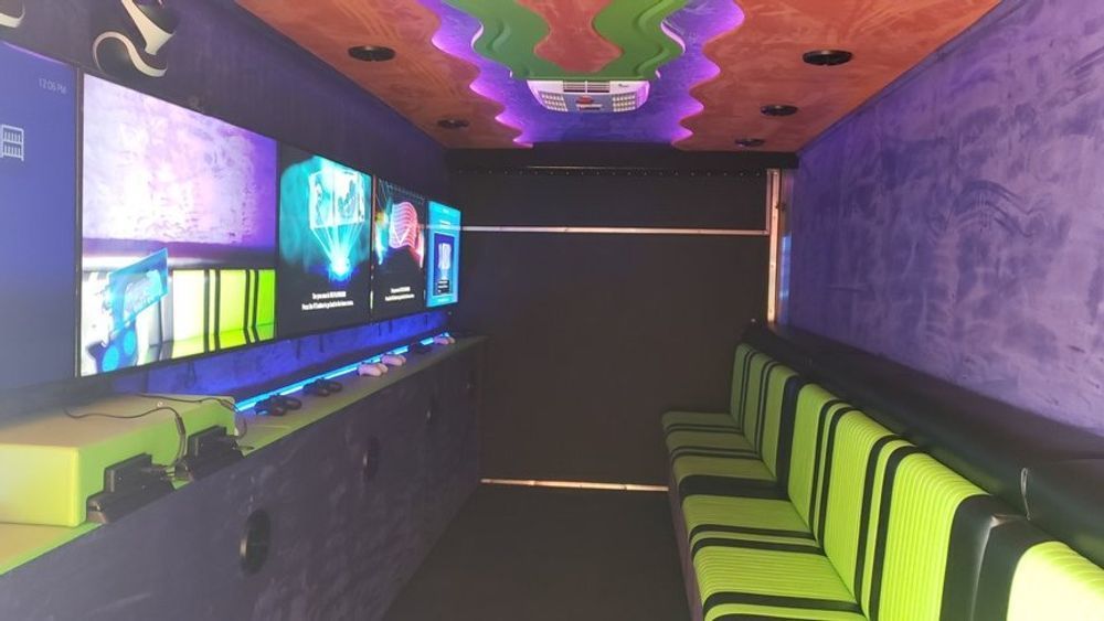 Interior of a gaming truck with multiple screens, neon green and black seating, and a colorful ceiling.