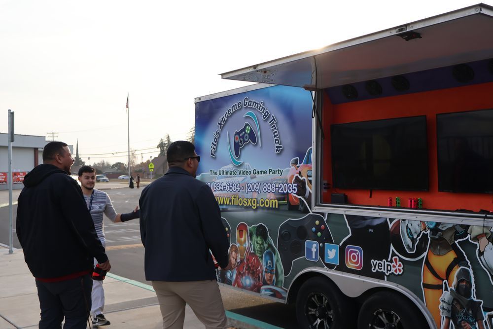 Men look at a mobile video game trailer, featuring game-themed graphics and multiple screens, parked outdoors.