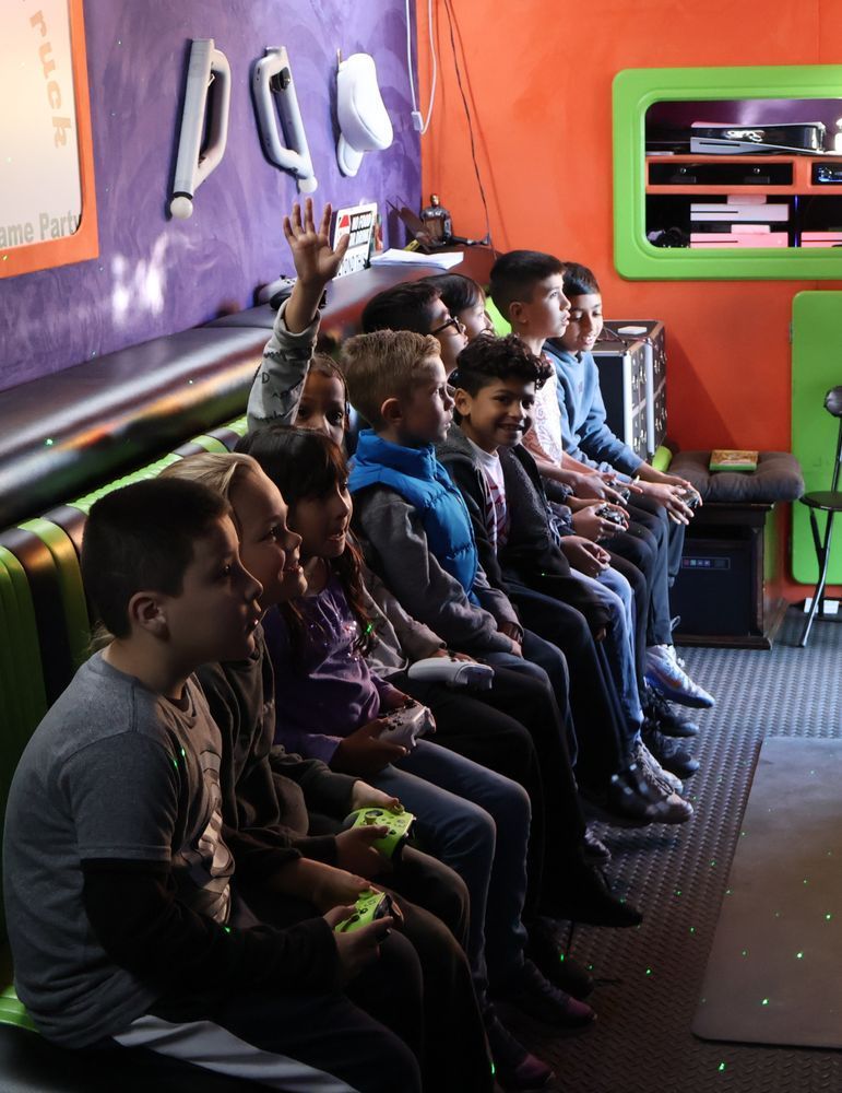 Group of children seated, one raising hand, in a colorful gaming room.