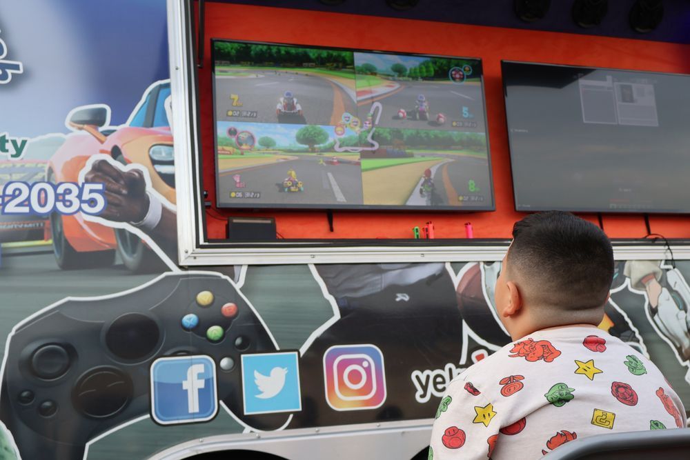 Boy watches a video game (Mario Kart) on a screen mounted on a game truck. Social media icons on the truck.