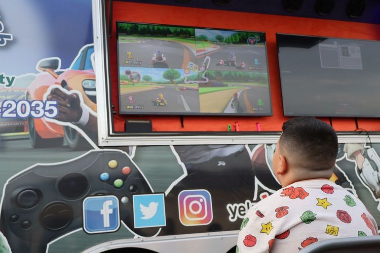 A child watches a video game on a screen in a gaming truck; social media icons on the side.