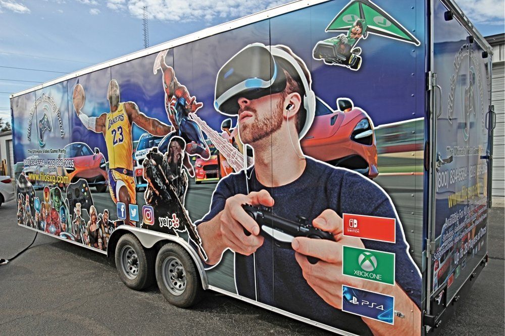 Gaming trailer with a man in VR gear. Colorful graphics include sports cars, a basketball player, and game console logos.