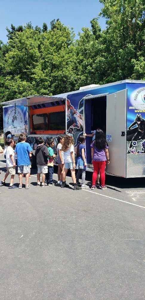 Children stand in line next to a truck with an open door on a sunny day. Green trees in the background.