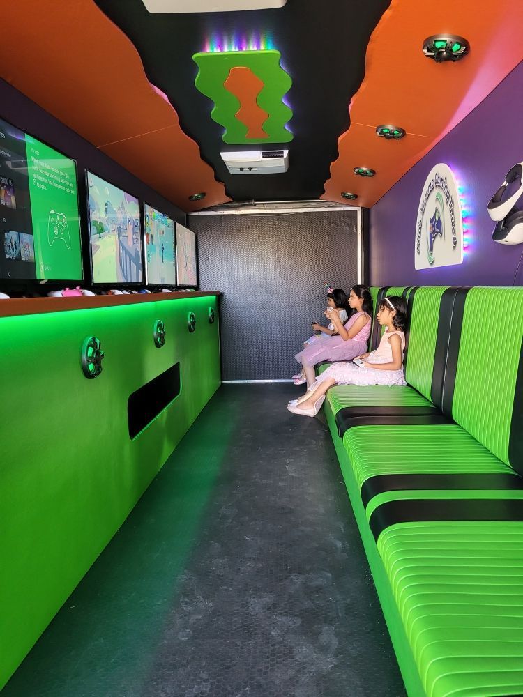 Three people in a bright green and orange gaming truck. They are seated on a green bench, facing screens, playing.