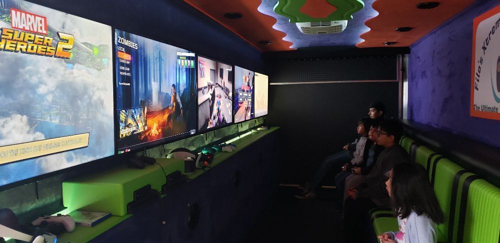 Inside a gaming truck, multiple screens display video games, and people sit on green benches.