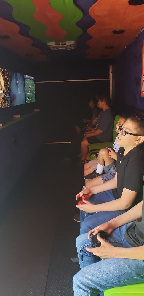 People play video games inside a colorful gaming truck, using controllers and watching screens.