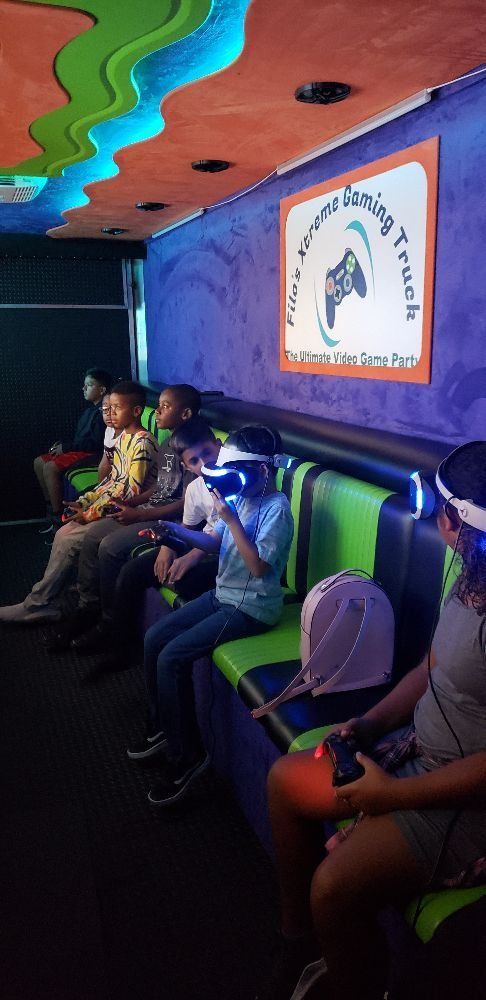 Children playing video games in a gaming truck. Some wear VR headsets and hold controllers. The interior has neon lighting and colorful decor.
