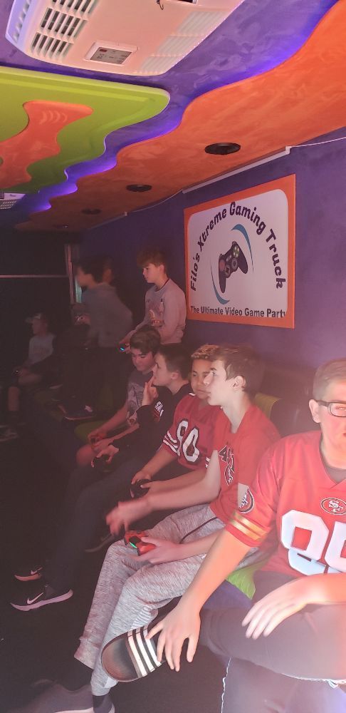 Inside a gaming truck, several people play video games while sitting on a long, cushioned seat. Orange, purple, and green decor.