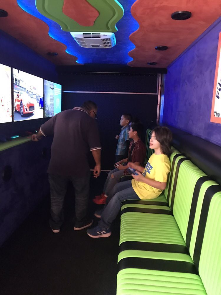 Inside a gaming truck, three children play video games. A man watches them. Neon green and black seating.