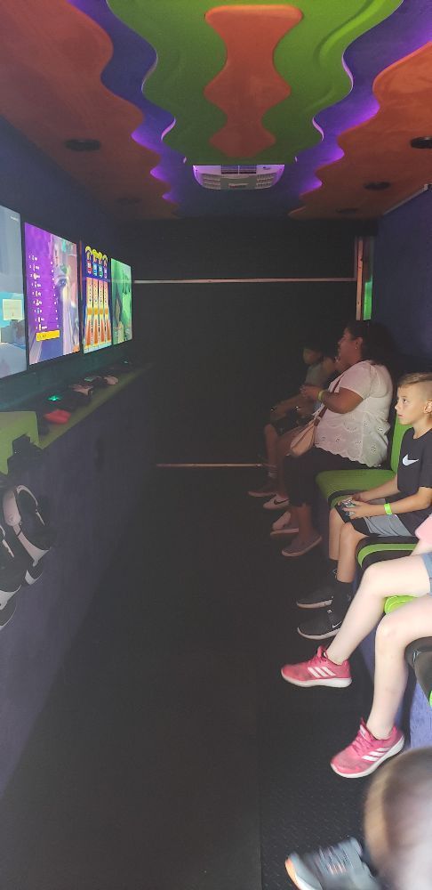 Inside a gaming trailer, people sit and play games. Colorful ceiling and screens.