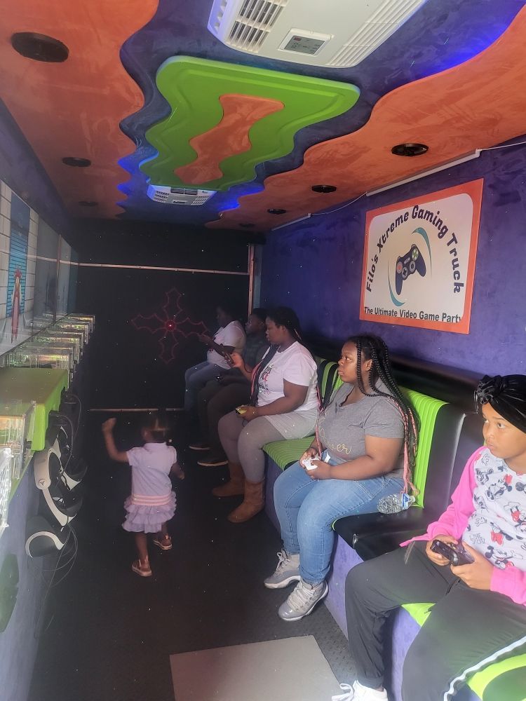 People playing video games inside a colorful gaming truck; a young child dances.